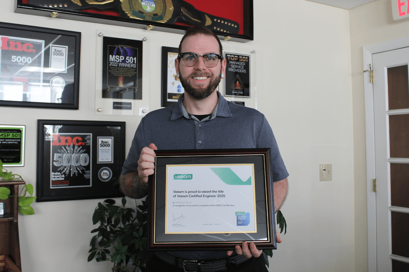 An ASG engineer holding his Veeam Certified Engineer 2025 certification by the ASG award wall